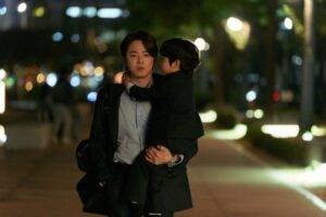 Photo of Jo Jung-seok as Han Jung-woo, holding his song in his arms with a serious expression on his face.